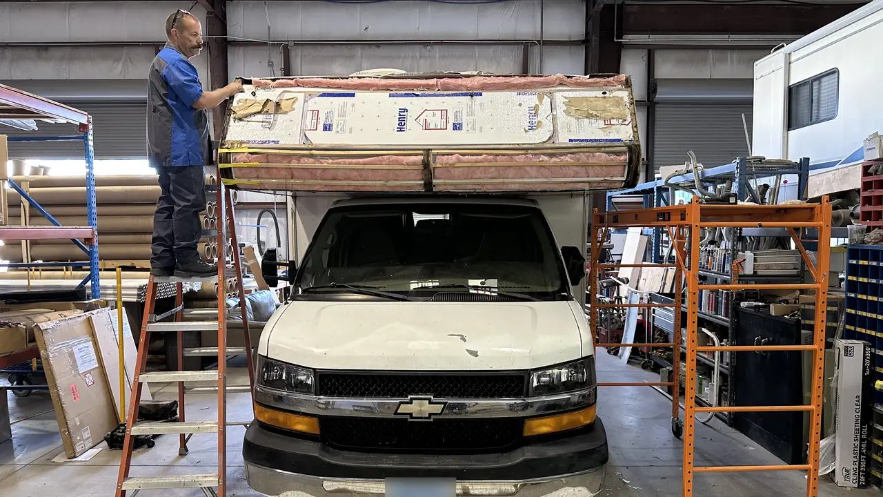 A technician on a ladder working on the cab-over roof section of a Class C motorhome inside the shop